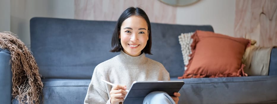 Portrait of asian woman with tablet, drawing, working on design project, holding pen, sitting in her living room, freelancing.