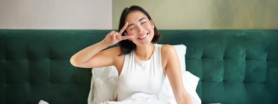 Positive asian woman lying in bed, showing peace sign, enjoys happy morning, waking up upbeat, staying in her bedroom.