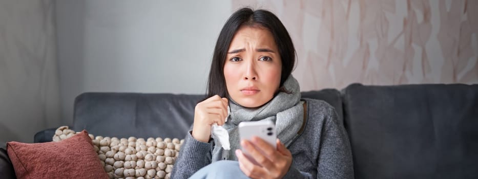 Asian girl with influenza, sits at home, cries and looks upset, holds smartphone, feels unwell and gloomy, wearing warm clothes.