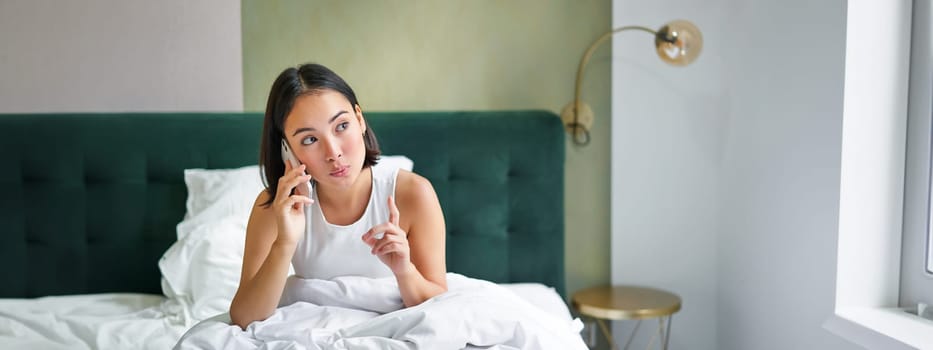 Beautiful young korean woman calling, having a phone call in bed, talking with friend while staying in bedroom.