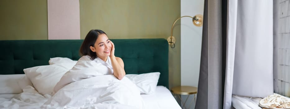 Beautiful sleepy asian girl wakes up in her bed with cozy white blankets, looking outside window with pleased smile. Lifestyle concept.