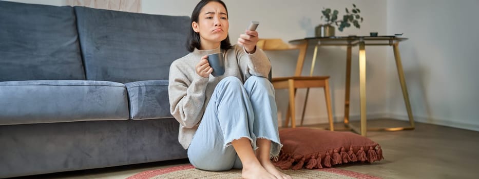 Portrait of girl watching television at home, sits on floor near sofa, holds remote and changes channels.