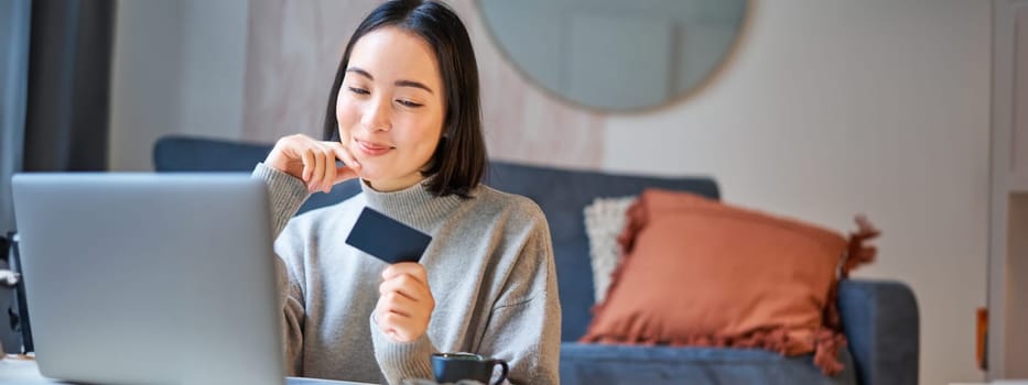 Happy smiling girl with credit card, paying her bills online on computer, doing shopping on her laptop, sitting at home.