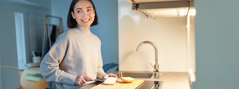 Cute asian woman making herself toast, cut loaf of bread, preparing sandwitch on kitchen.