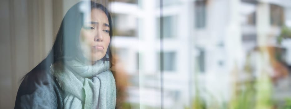 Portrait of sad ill korean woman looking out of the window, staying at home due to coronavirus, being sick and cant leave house.