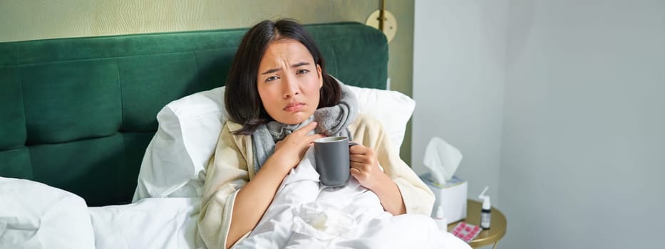 Health, flu and people concept. Portrait of korean woman feeling sick, catch a cold, staying in bed unwell, drinking hot tea.