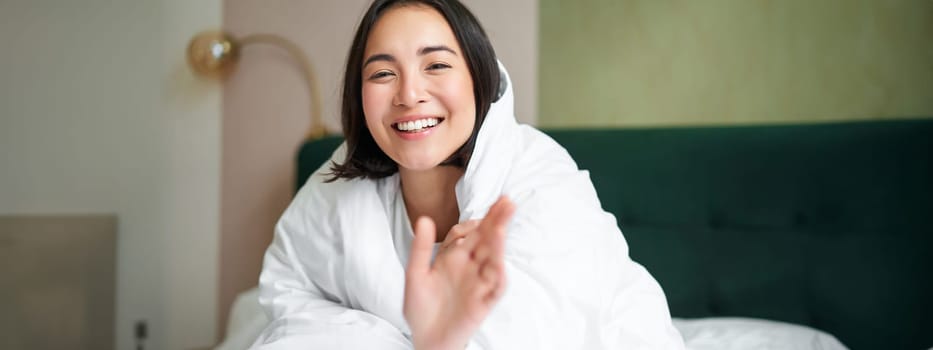 Happy beautiful asian woman covered with blanket, sits on bed in warm duvet, tries to reach camera with a hand, smiling playfully. Hotel bedroom, comfort and apartments concept.