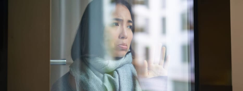 Portrait of sad ill korean woman looking out of the window, staying at home due to coronavirus, being sick and cant leave house.