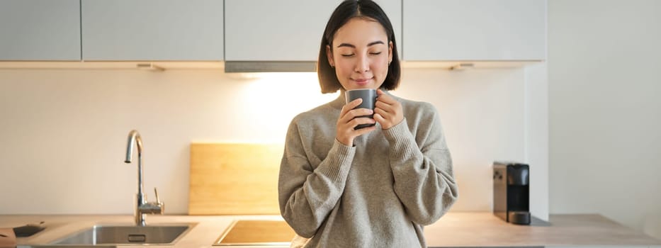 Portrait of smiling asian woman standing on her kitchen, drinking coffee and looking at camera, concept of cozy and comfortable home.