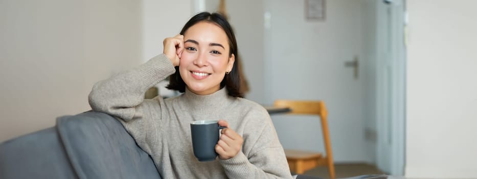 Smiling asian woman sitting at home with cup of coffee, relaxing and feeling warmth, looking outside window, resting on sofa in living room.