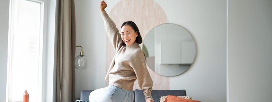 Cute excited asian woman dancing, raising hands up in triumph, celebrating and singing, standing at home in warm jamper.