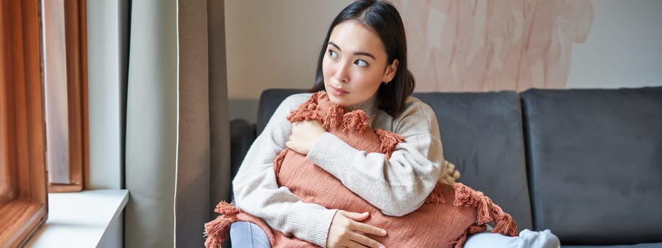 Portrait of asian woman feeling cozy in her home, hugging pillow on sofa and looking thoughtful, smiling.