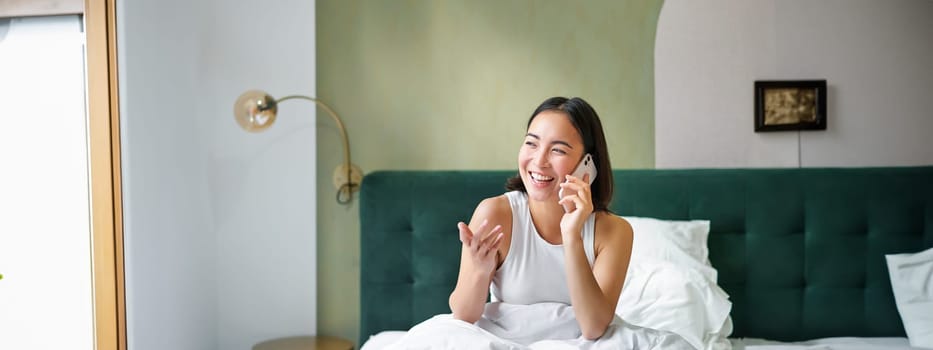 Beautiful smiling asian girl talking on mobile phone, lying in bed with pleased happy face, speaking to someone on telephone.