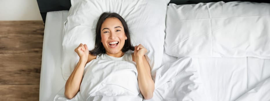 Top view of enthusiastic asian girl cheering, lying in her bed and shouting with joy and excitement, triumphing and smiling, enjoy staying in her bedroom.