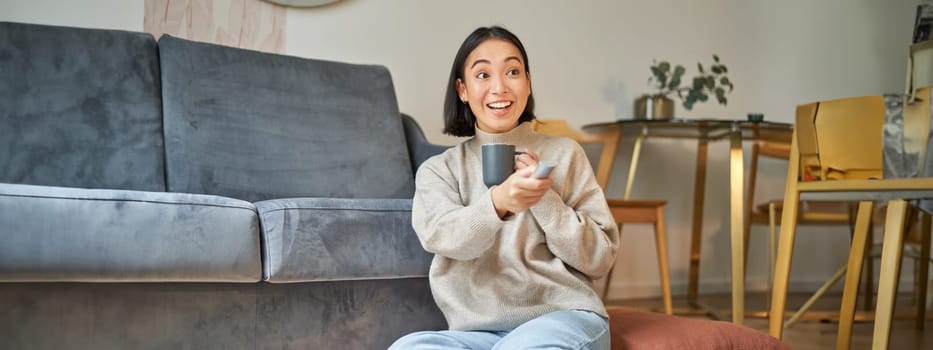 Portrait of smiling young woman resting at home near tv, watching television sitting on floor and drinking coffee from cup.