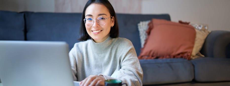 Self-employed young smiling woman, freelancer staying at home, working on remote from laptop, wearing glasses, sitting in living room.