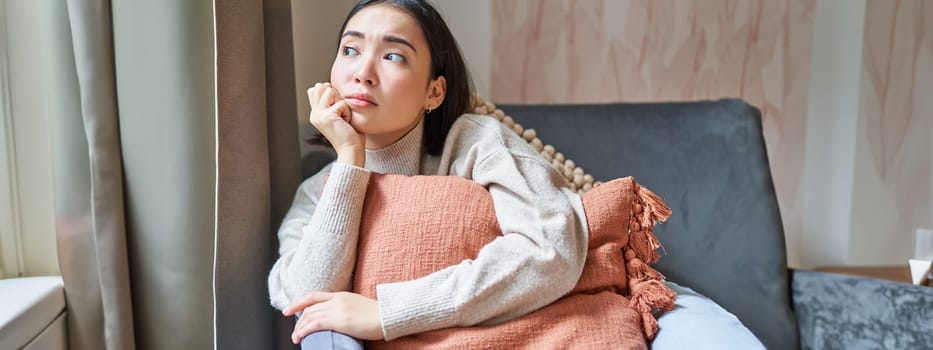 Women and wellbeing concept. Portrait of thoughtful asian woman sitting on sofa with pillow, looking aside with thinking concentrated face.