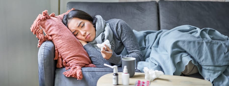 Sick sad korean woman lying on sofa, feeling unwell, catching cold, flu and temperature, looking upset, taking medication.