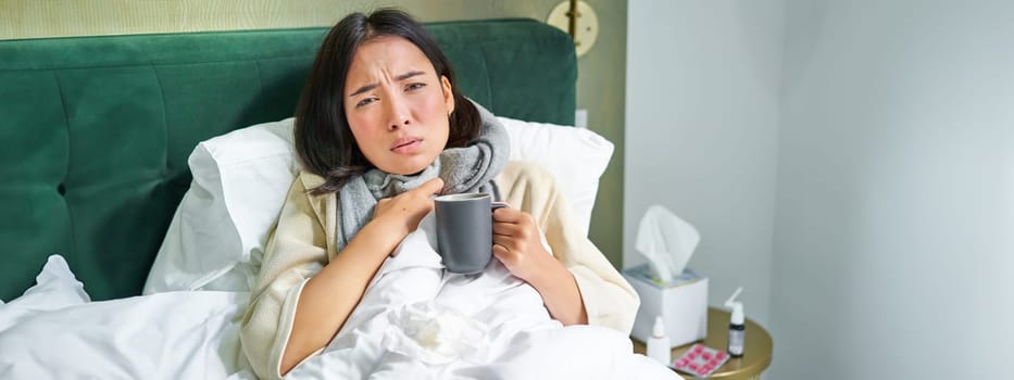 Portrait of couching asian girl in bed, feeling sick, catching cold and staying at home, looking unhealthy, drinking hot tea.