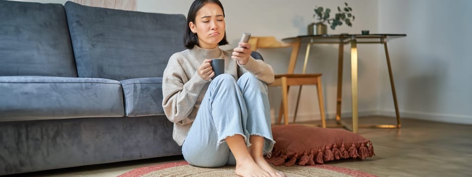 Portrait of young woman with remote, watching tv, switching chanels on television, sitting on floor near sofa and relaxing.