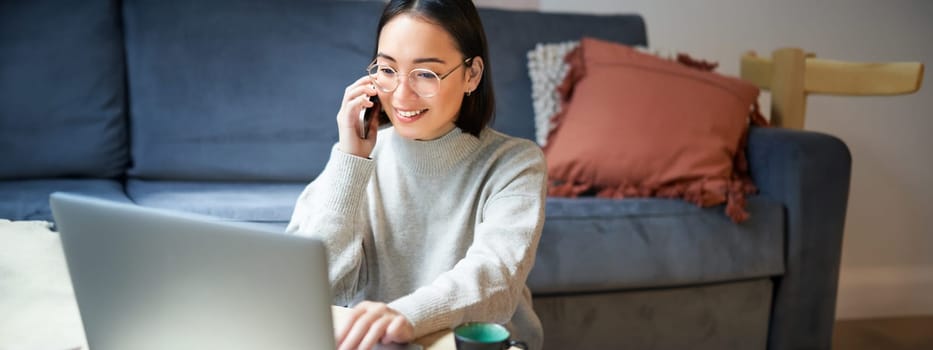 Portrait of smiling asian woman making phone call, working on laptop and having conversation on smartphone.