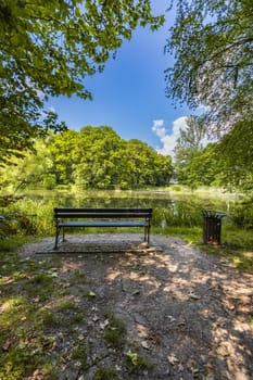 Old wooden bench in beautiful and colorful park at sunny morning with blue sky with few clouds beautifully reflecting in big silent lake like in mirror