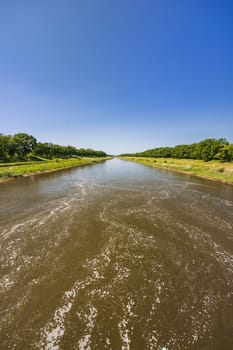 Beautiful landscape of long dirty river with green trees and bushes on both sides seen from bridge over water dam and water level