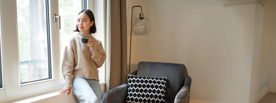 Vertical shot of beautiful woman sitting in her house, looking outside window and drinking coffee.