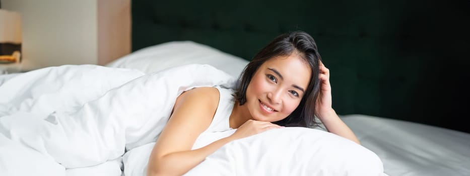 Portrait of young korean woman smiling, lying in bed on pillow, posing in her bedroom, looking relaxed and feeling comfortable.