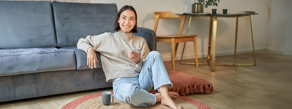 Portrait of stylish korean woman sits on floor with smartphone, using mobile phone, smiling pleased, concept of staying at home and relaxation.