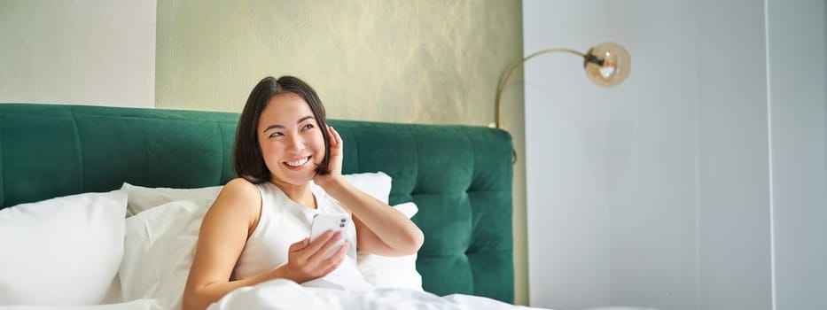 Cute korean girl in bed, holding smartphone, feeling happy and pleased, spending morning in bed, enjoying surfing net on mobile phone.