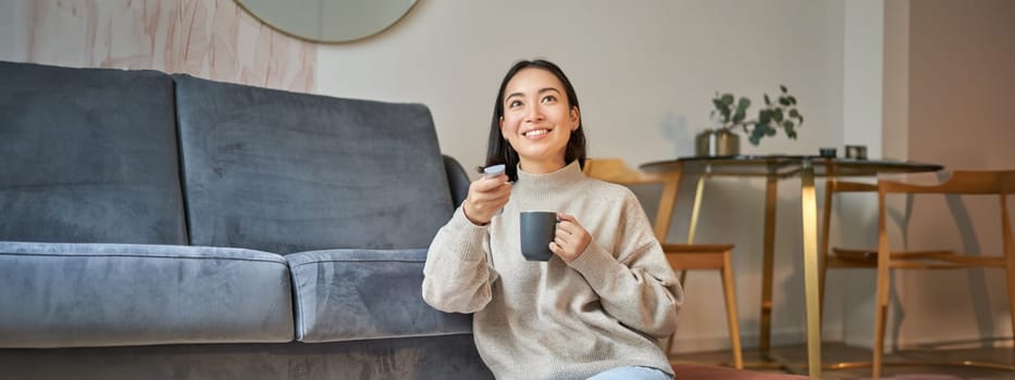 Portrait of smiling korean woman sitting near tv, holding remote and switching channels while drinking hot coffee.