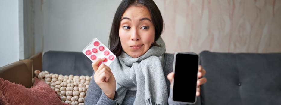 Happy asian woman catching cold, showing smartphone screen and pills, recommending online doctor app, GP or medical clinic on phone, holding drugs, staying at home with flu,