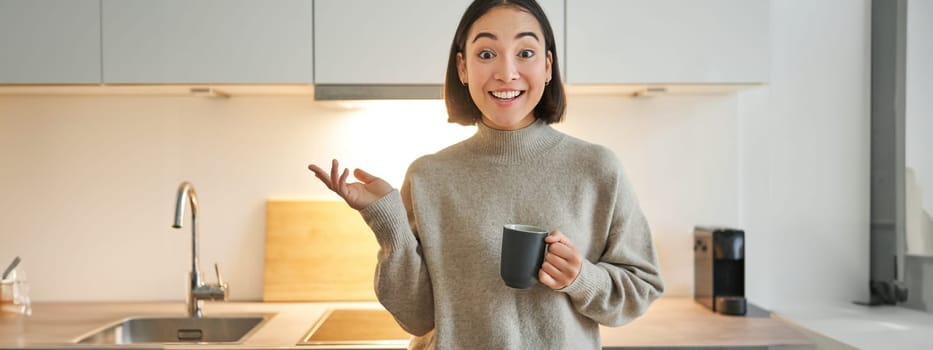 Portrait of stylish asian woman standing in kitchen with mug, drinking coffee, explaining smth, showing banner or advertisement.