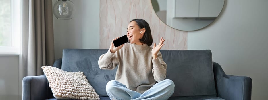 Portrait of carefree asian girl sits at home, listens to music in headphones and sings at smartphone, holds mobile phone like microphone.