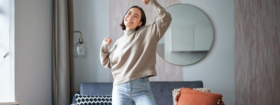 Portrait of happy asian woman dancing, rejoicing and triumphing, feeling upbeat at home. Copy space