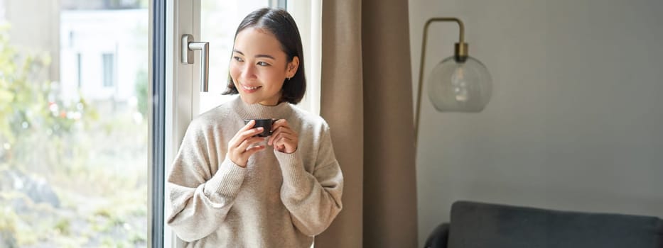 Portrait of beautiful asian woman sitting at home with cup of coffee, enjoying her espresso while looking outside window at passerby, smiling happily.