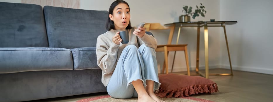 Portrait of girl watching television at home, sits on floor near sofa, holds remote and changes channels.