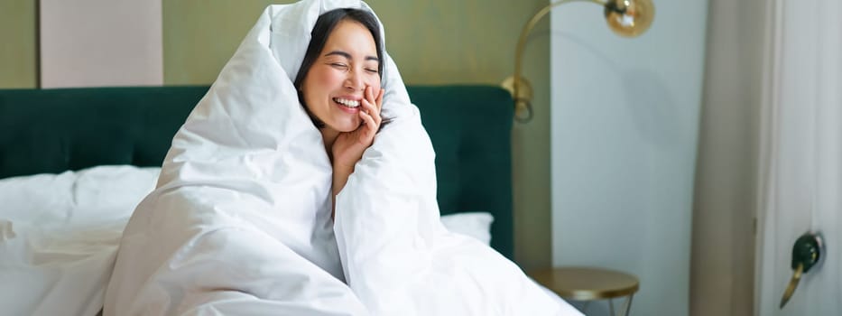 Beautiful asian woman sitting on bed, covered in blankets and duvet, laughing and smiling, enjoying weekend in bedroom.