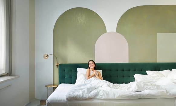 Vertical shot of beautiful asian girl lying in bedroom with sophisticated interrior, smiling and looking happy in morning, spending time in bed.