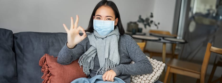 Covid-19 and health concept. Young asian woman in medical face mask, feels sick and unwell, catching flu, protecting others from influenza, sitting in living room covered with blanket.