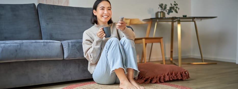 Portrait of girl watching television at home, sits on floor near sofa, holds remote and changes channels.