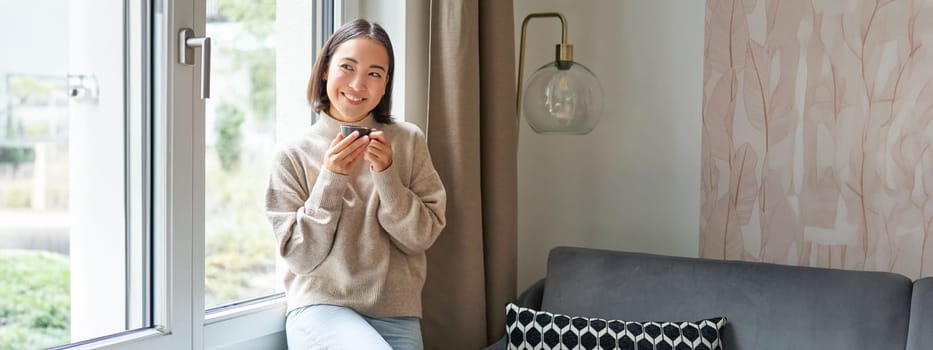 Beautiful young asian woman sitting near window and drinking her coffee, holding espresso cup and looking outside with relaxed, smiling face expression.