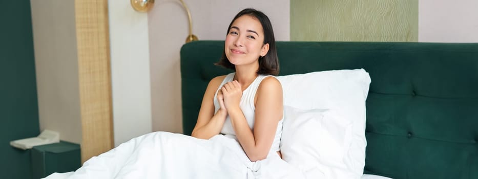 Portrait of excited, smiling asian woman sitting in her bed in morning, daydreaming, looking hopeful.