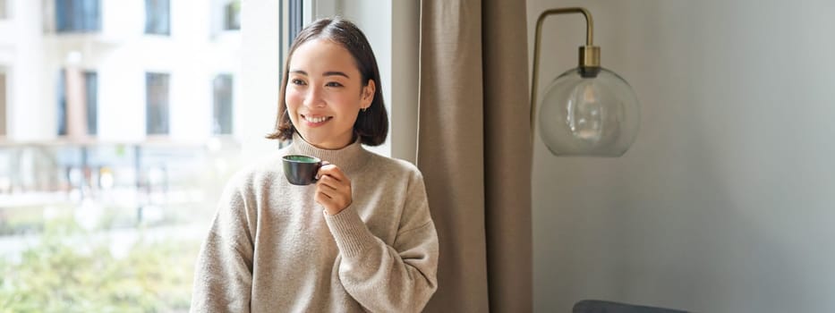 Portrait of beautiful asian woman sitting at home with cup of coffee, enjoying her espresso while looking outside window at passerby, smiling happily.