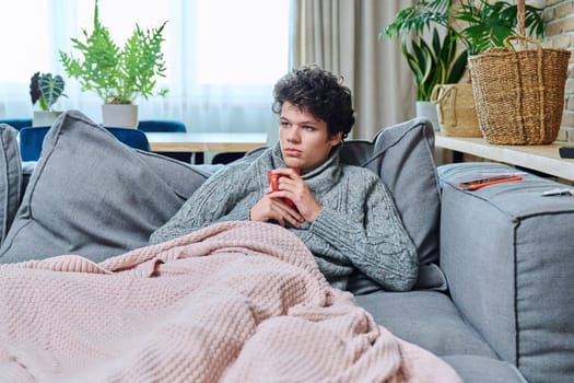 Young sick male in warm sweater with mug of hot drink under plaid relaxing at home on sofa in living room, with signs of respiratory diseases. Cold flu season health lifestyle youth