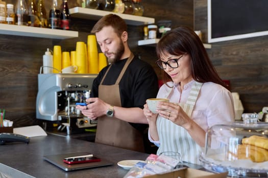 Team, partners, young male and mature woman talking working using phone standing behind bar in coffee shop. Team, small business, work, staff, cafe cafeteria restaurant, entrepreneurship concept