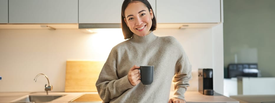 Portrait of smiling asian woman standing on her kitchen, drinking coffee and looking at camera, concept of cozy and comfortable home.