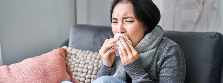 Close up of young korean woman staying at home with cold, sneezing in napking, has runny nose, concept of illness, health and influenza.