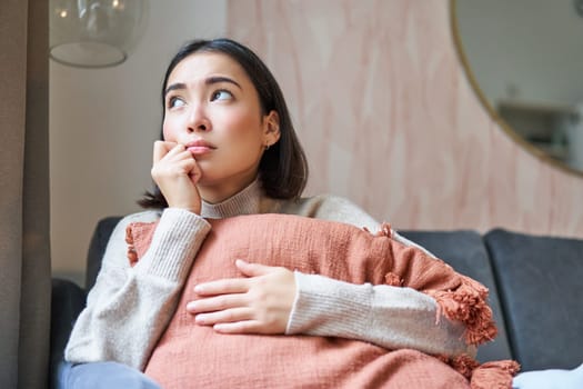 Women and wellbeing concept. Portrait of sad and gloomy asian woman thinking of something unhappy, hugging pillow on sofa and expressing distress.